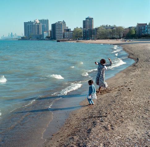 A woman in a black and white dress is about to skip a stone at a beach in the afternoon light with her young daughter.