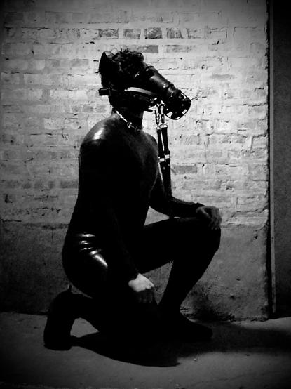The raw, evocative image showcases a striking monochromatic composition featuring a person kneeling in a dimly lit space. They are dressed in a sleek, form-fitting silicone bodysuit that gleams under the dim light, emphasizing the contours of their body. The suit’s texture adds a futuristic, almost otherworldly quality to the scene.

The focal point of the image is the intricate leather hood they wear, designed in the style of equine tack. The hood features a muzzle-like structure that wraps snugly around their face, complete with straps and buckles that suggest both restraint and control. A metal ring at the front connects to a lead or rein, reinforcing the human-horse dynamic central to this visual narrative. The attention to detail in the hood’s craftsmanship—its stitching, hardware, and design—enhances its aesthetic and symbolic impact.

The background is stark and industrial: a rough brick wall bathed in shadow. This setting contrasts sharply with the polished appearance of the figure, creating a dramatic interplay between texture and tone. The dramatic lighting enhances the mood, casting deep shadows that amplify the intensity and intimacy of the moment, making the image  deeply atmospheric.