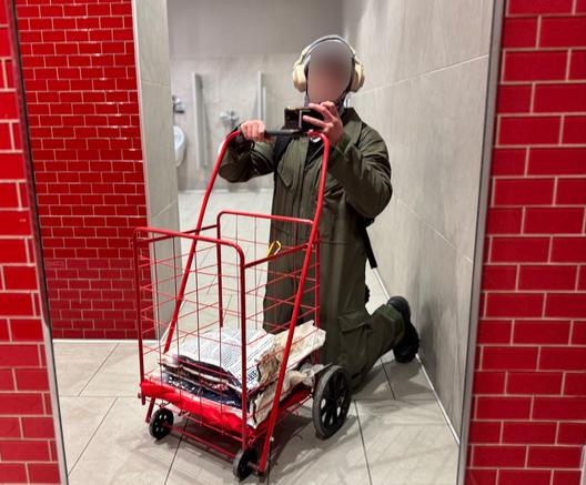 The image situates an individual in a brightly lit, utilitarian environment—a public restroom with bold red tiles that dominate the scene. The olive-green work suit they wear is functional and unassuming, designed for practicality rather than aesthetics. The red utility cart, loaded with neatly stacked goods, becomes an extension of their role. Its vibrant color mirrors the tiles, creating a visual harmony that underscores their integration into this environment.

The subject assumes a kneeling posture that conveys humility and dedication but also hints at a deeper narrative of service. The heavy-duty headphones serve as both a barrier and a tool—perhaps blocking out distractions while signaling focus on the task at hand, or playing looped hypnosis recordings. This image captures the essence of submission as a deliberate act of purpose, where even mundane chores can be imbued with dignity, meaning, and care.