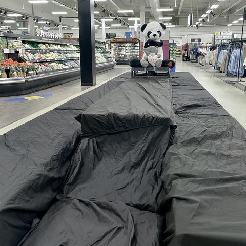 A grocery store aisle featuring a large, draped black tarp with a stuffed panda bear and a smaller stuffed animal sitting on a cart. The background includes shelves of fresh produce and clothing racks.