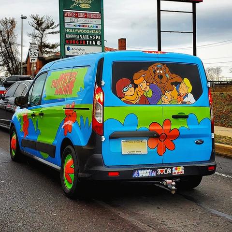 A van painted to look like the famous cartoon vehicle. Blue with splashes of green with red lettering. Scooby Doo, Shaggy, Daphne, Velma, and Fred are painted on the back window as if they're looking out from the van.