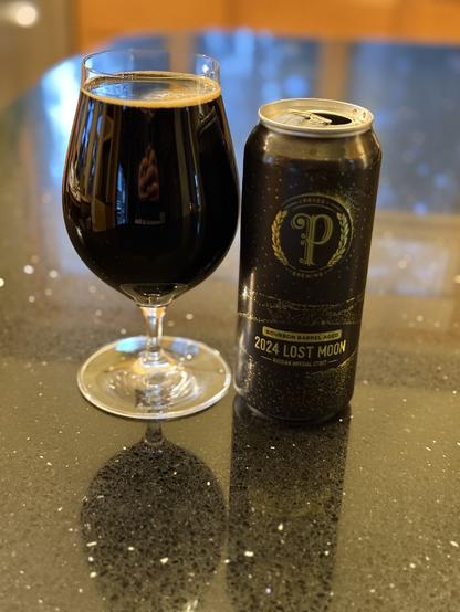 A tall snifter full of dark beer. The black pint can has highlights in gold, a star field and large scripted “P” surrounded by a fleur de lea