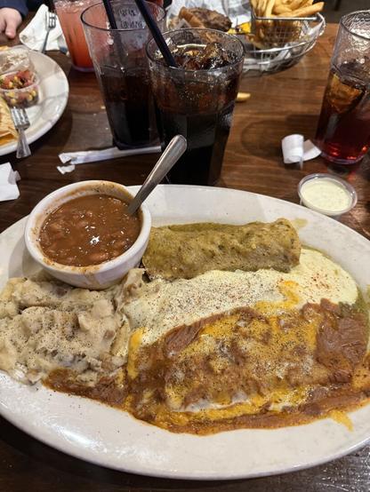 The image shows a plate of food on a wooden table at a restaurant. The plate contains a serving of mashed potatoes sprinkled with black pepper, a small bowl of baked beans with a spoon, and an enchilada covered in a mix of green and red sauces with melted cheese on top. In the background, there are two glasses filled with a dark soda and ice, each with a straw. There is also a basket of fries and a burger partially visible, along with a small container of a creamy dressing or sauce. Other plates with food and drinks can be seen around the table.