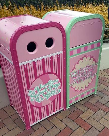 Two colorful trash cans designed with a candy theme. The left can is pink with a circular design and two holes for recycling, labeled "Vanellope's Sweet Pop World." The right bin is pink and green, featuring a decorative border and the same branding.