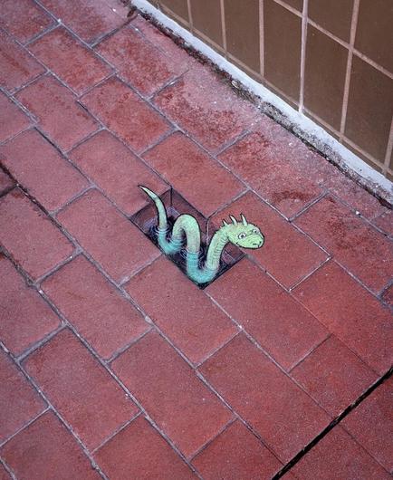 Streetart. A green snake (called Roger) and a "hole" have been drawn in chalk on a red brick sidewalk. Roger meanders through the brick-sized hole filled with painted water and looks around expectantly. Title: "Roger is secretly fond of attention and the Loch Ness Monster's most embarrassing cousin."