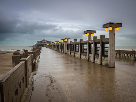 A medium shot shows a pier on a beach, under an overcast sky. The pier is made of concrete and has a low wall on either side. At regular intervals along the pier are concrete columns with a small window between each column. On top of each column is a square with four orange spherical lights. In the distance is a building with a tower on top, at the end of the pier. The beach is visible on either side of the pier, and the sea in the background. The sky is gray and cloudy. The pier is wet, reflecting the sky.