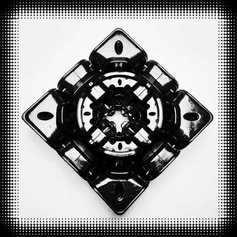 Foto en perspectiva centila de una capa independiente del cubo de Rubik sin su centro. Son un total de ocho piezas de plástico negro.
Así se puede ver que el interior de un cubo está lleno de recovecos, formas redondeadas y huecos para que fluya el lubricante (razón por la que brillen tanto).
La foto tiene un marco negro formado por puntos que se "diluye" a medida que los círculos se hacen más pequeños.