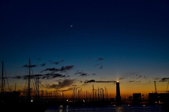 bild im querformat zur blauen stunde:
bild im hafen, in rechter unteren bildecke steht ein lecuhtturm, der gerade licht in richtung kamera wirft. der hafen ist voll mit segelyachten.
Der himmel färbt sich vom tiefen orange über sichtem gelb/blau ins dunkle blau des nachthimmel. am himmel hänge eine dünne mondsichel