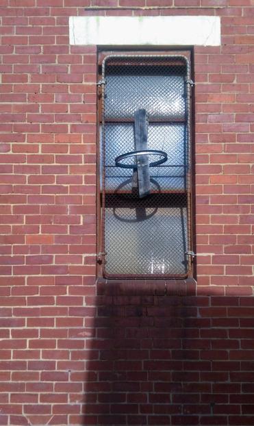 A makeshift basketball hoop made of a round metal rim nailed to a vertical plank of old, grey wood is mounted on the wire security mesh covering a tall window set into the brick wall of an industrial building. Harsh sunlight glints off the window, and the bricks below the window are in deep shadow.