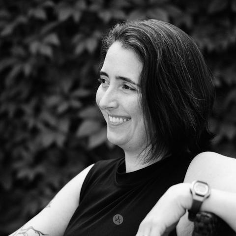 Black and white head shot of Hilary. The photo is taken from the side. She is sitting in a chair, leaning her left arm on the armrest. in the foreground. She's smiling and looking to the left side oft the photo. The background is blurry.