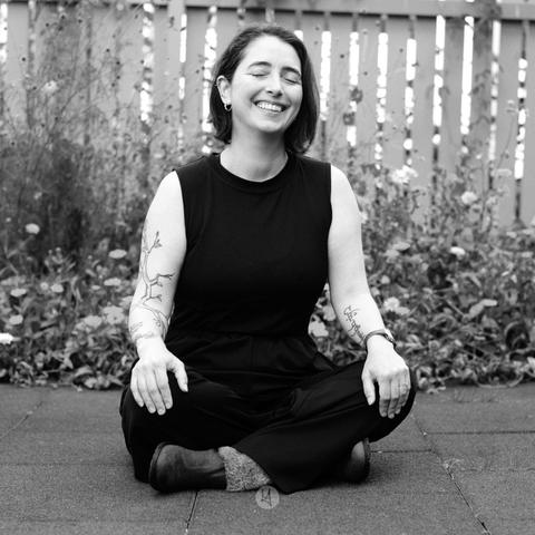 Black and white photo of Hilary sitting cross-legged on a mat of a playground faced directly to the camera. Her eyes are closed and she's is smiling.