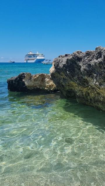 Picture of the ocean and blue sky with a cruise ship in the distance. Closer is located a big rock formation next to the shore.