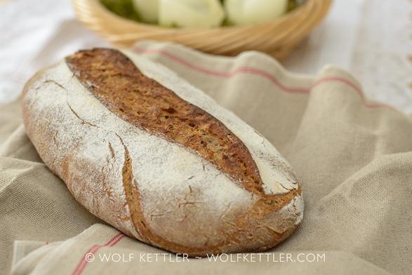 The photograph shows an oblong loaf of rustic bread with scoring running along the centre. The bread is displayed on a piece of baker’s linen.