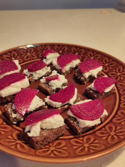 A brown plate with a selection of small sandwiches: brown bread, white cream cheese and purple slices of beetroot on top