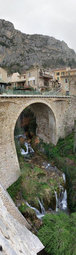 Historic stone bridge in Moustiers-Sainte-Marie with a waterfall cascading beneath, set against a backdrop of traditional hillside architecture.