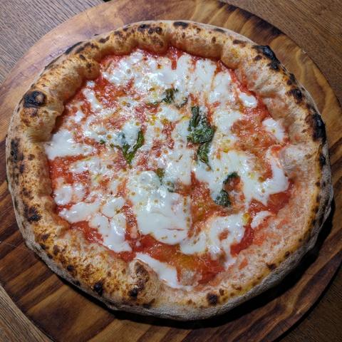 Eine Pizza Margherita auf einem Holzbrett von oben fotografiert. Die Rand ist luftig und dick, die Pizza mit Tomatensoße, Käse und Basilukum belegt.