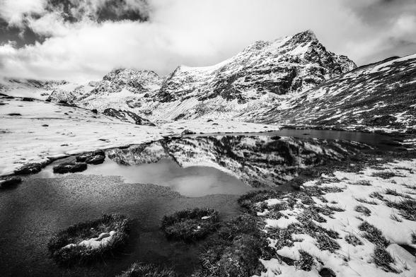Photo d'un paysage de montagne en noir et blanc.
Des montagnes saupoudrées de neige se reflètent dans un petit lac de montagne. La neige ne recouvre pas tout le sol, on distingue des zones herbeuses.
Au premier plan il y a deux ronds d'herbe qui émergent de l'eau, avec de petites taches de neige dedans.
Le ciel est nuageux.
-
Photo of a mountain landscape in black and white.
Snow-dusted mountains are reflected in a small mountain lake. The snow doesn't cover the whole ground, and grassy areas can be made out.
In the foreground there are two grass rings emerging from the water, with small patches of snow in them.
The sky is cloudy.