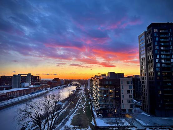 he image depicts a scenic view of a river at sunset, with vibrant colors in the sky ranging from orange to purple. On either side of the river, there are modern buildings, some of which have lights on, indicating activity inside. The foreground features a snowy landscape along the riverbank, adding to the winter atmosphere. The overall scene conveys a peaceful yet dynamic urban environment during twilight.