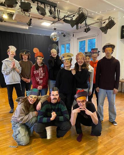 Eine Gruppe von 12 Jugendlichen steht in einem Raum mit Holzboden. An der Decke befinden sich Bühnenlichter und eine Discokugel. Die Jugendlichen tragen Masken, die Gesichter älterer Menschen mit verschiedenen Hüten darstellen. Einige der Jugendlichen stehen, während andere knien. Sie tragen verschiedene Arten von Freizeitkleidung. Die Stimmung ist fröhlich und verspielt.