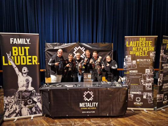 Metalityheads aus den Chapters Frankfurt und Karlsruhe beim Gruppenbild. Sie stehen am Infostand beim Papa Roach Konzert in Frankfurt/Main. Vor der Gruppe befindet sich ein Tisch mit Metality-Flyern, das Metality-Logo auf einem großen Banner befindet sich an der Wand hinter den Personen und ein kleineres Banner mit Metality-Logo ist am Tisch befestigt. Neben der Gruppe stehen Rollups mit den Schriftzügen "Family. But Louder." und "Das lauteste Netzwerk der Welt."