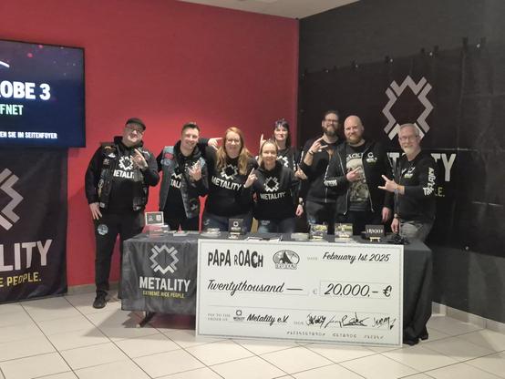 Metalityheads aus den Chapters Düsseldorf und Ruhrpott  am Infostand vor dem Papa Roach- Konzert Konzert in Düsseldorf. Im Hintergrund der Gruppe befindet sich das Metality-Logo auf einem großen Banner. VOr der Gruppe  ist ein Tisch mit Metality-Flyern aufgestellt. Am Tisch leht ein großer Spendenscheck über 20.000 €, den die Crew von Papa Roch überreicht bekam.