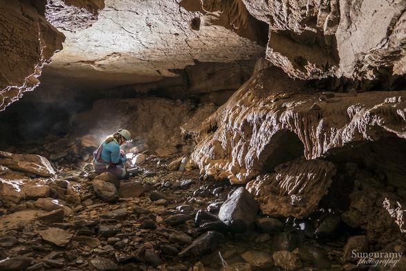 A backlit cave scene with a long haired person in purple and teal sitting left center, holding a small net in hand, looking intently at it. The ground is cobble covered rocky creek inside the cave, there is obviously water around. On the right is some nice flowstone formation which kinda is like if a waterfall was made of rock.