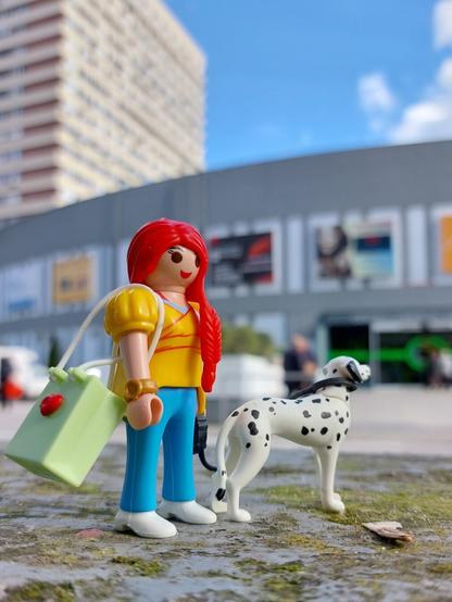 Menina playmobil de cabelo ruivo comprido, calças azuis e camisa amarela, saco de compras verde segura pela trela um cão dalmata que olha para um shopping verdadeiro