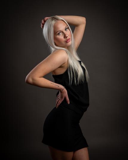 The image shows a person with long, light-colored hair posed against a plain, dark background. They are wearing a black sleeveless outfit and have one hand on their hip while the other is resting on their head. The lighting highlights their features, giving the photograph a professional studio look.