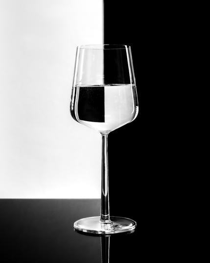 The image shows a wine glass filled halfway with water. The background is split into two contrasting halves: white on the left and black on the right. The glass is positioned so that it visually interacts with the divided background, creating an interesting optical effect.