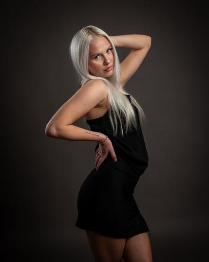 A young blonde woman confidently poses in a studio against a dark background, wearing a stylish black dress. The lighting enhances her facial features and the shine of her hair.