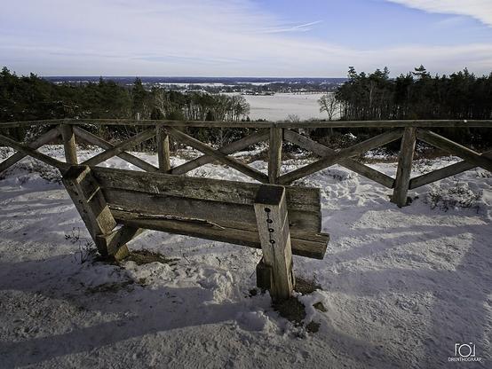 Viewpoint on a hill. A wouden bench and a wouden fence keep the visitor from falling of the edge.  The surroundings are covert in snow.