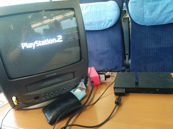 Samsung CRT next to a PlayStation 2 slim on a table inside an ICE train. The CRT screen displays the PlayStation 2 logo.