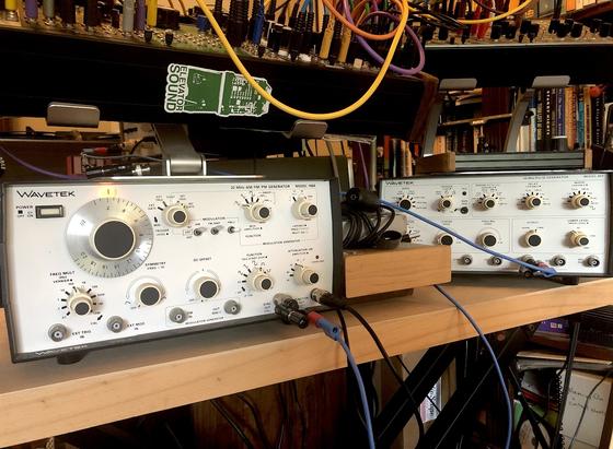 Photo of two pieces of equipment, both made by Wavetek. Both are rectangular with white faces and lots of dials. Also bnc connections for ins and outs. There's a blue cable running from one to the other, and the left one has a black cable plugged into it's output. Other pieces of equipment can be seen above.