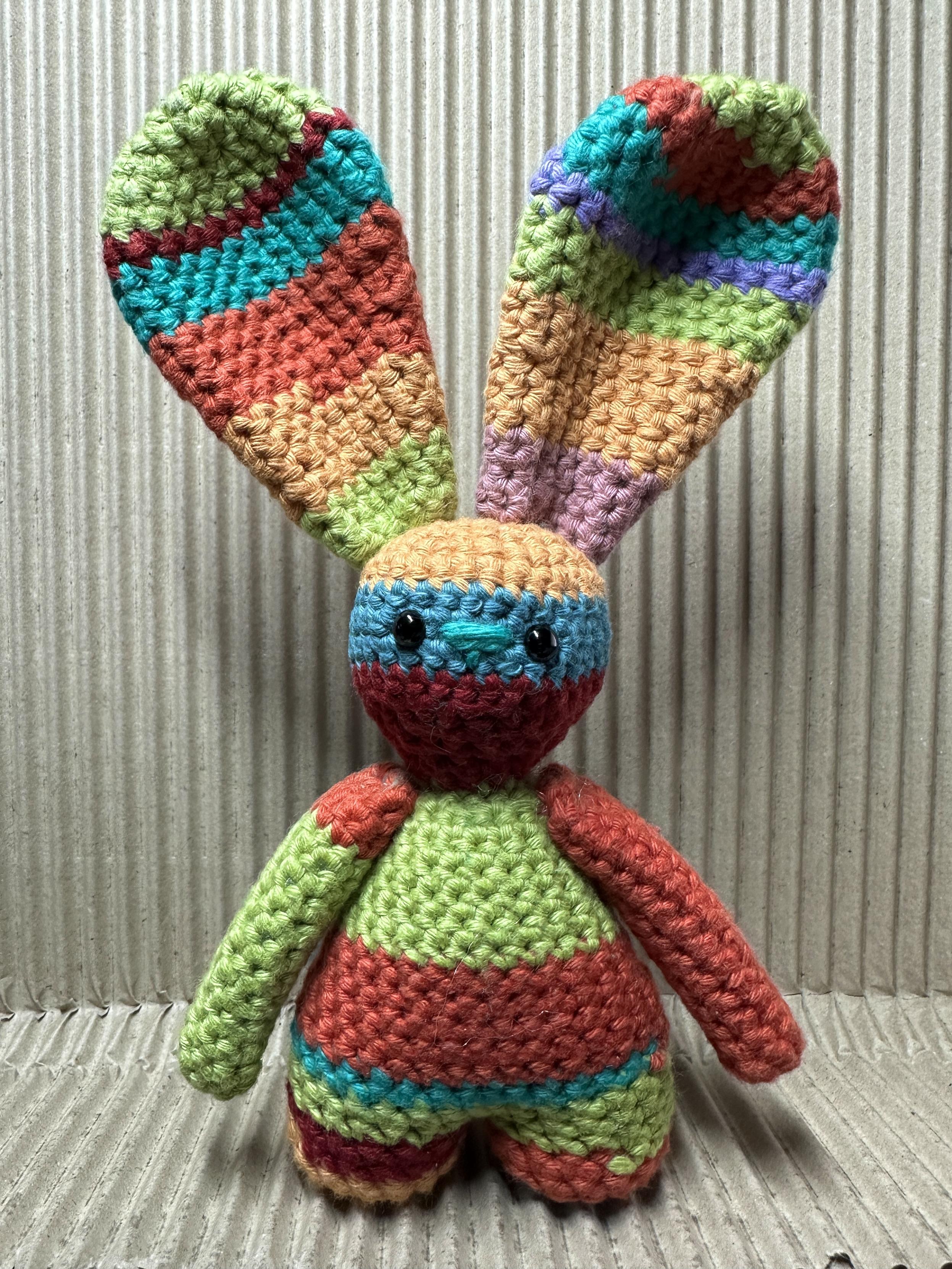 Small crocheted rabbit. It’s made from lots of different coloured pieces of cotton yarn. It’s easy are far to big for it and its legs are very short