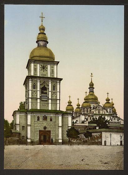 The image depicts a historical photograph of St. Michael's Monastery in Kiev, Russia (now Ukraine). The monastery is captured with its iconic baroque architecture prominently displayed. It features several onion-shaped domes painted in green and gold hues, which are characteristic of Ukrainian Orthodox churches.

In the foreground, there’s a large bell tower with intricate geometric patterns on its facade, topped by a golden dome containing a cross at its pinnacle. The photograph appears to be taken during dusk or dawn due to the soft lighting conditions; this gives an ethereal glow to the scene.

The surrounding area is open and spacious, possibly indicating that it was situated in relatively less densely built urban areas of Kiev around that period. There are some trees visible on the left side, adding a touch of greenery to the otherwise stony pavement or plaza where the monastery stands.

Overall, this image provides an insight into the architectural beauty of historical Eastern European churches and their surroundings from approximately late 19th-century Russia (Ukraine).