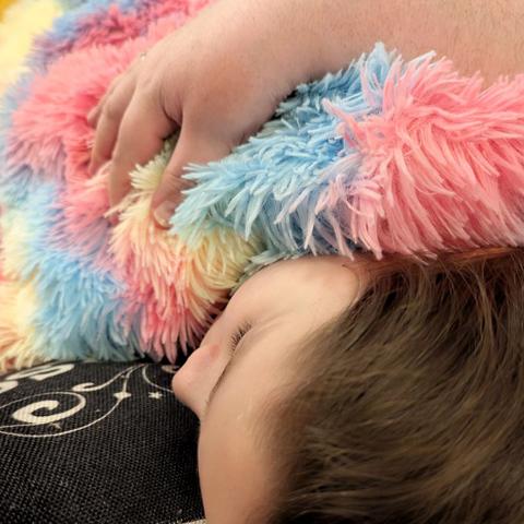 Liegender Kinderkopf mit braunen Haaren auf Kissen, das Gesicht ist aus dem Winkel nicht wirklich erkennbar, die Augen sind geschlossen. Eine gelb-rosa-blaue Decke mit langem Monsterflauschfell bedeckt das Kind. Eine elterliche Hand liegt auf Rücken und Schulter und hält das schlafende Kind fest und sicher.