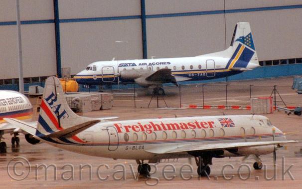 Side view of a bare metal finish, 4 propellor-engined airliner parked facing to the right and slightly away from the camera.
There is a thin, blue and red stripe running along the body, rising up into the tail and getting wider.
There are large red "Freightmaster" titles on the upper fuselage, and blue "BAF" titles on the tail.
The registration "G-BLOA" is on the lower rear fuselage.
A white, twin engined jet airliner is partially visible on the left of the frame, while a white and blue, twin propellor-engined airliner is parked in front of the grey doors of a large hangar in the background.