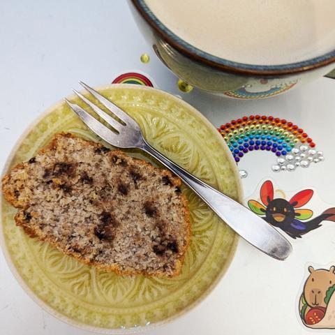 Limonengelber, kleine Kuchenteller, darauf ein Stück Rührkuchenin Kastenform. Beigefarben mit Schokostückchen. Sieht nicht nach viel aus, ist aber lecker. Daneben eine Tasse mit Milchkaffee.