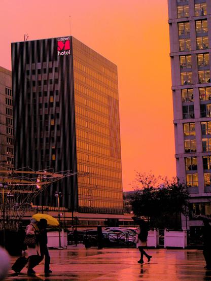 La photographie représente une scène urbaine au coucher du soleil, avec un bâtiment portant le logo « citizenM hotel ». Le ciel a une teinte orange chaude et des personnes marchent au premier plan, certaines portant des parapluies. Les bâtiments reflètent la couleur du coucher de soleil, créant une atmosphère vibrante.