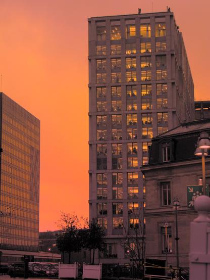 La photographie représente une scène urbaine au coucher du soleil. On y voit des immeubles de bureaux modernes dont l'intérieur est illuminé, avec les mêmes teintes chaudes du ciel au coucher du soleil. Le ciel est d'une couleur orange frappante, ce qui renforce l'atmosphère générale. Au premier plan, des arbres et quelques lampadaires contribuent à créer un paysage urbain.