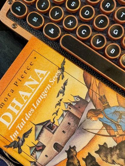 Oranges Buchcover mit einem gezeichneten Mädchen mit Bogen, Burg und Wölfen. Es ist das Kinderbuch "Dhana im Tal des langen Sees". Daneben eine Retro-Tastatur in Kupfer und Schwarz.