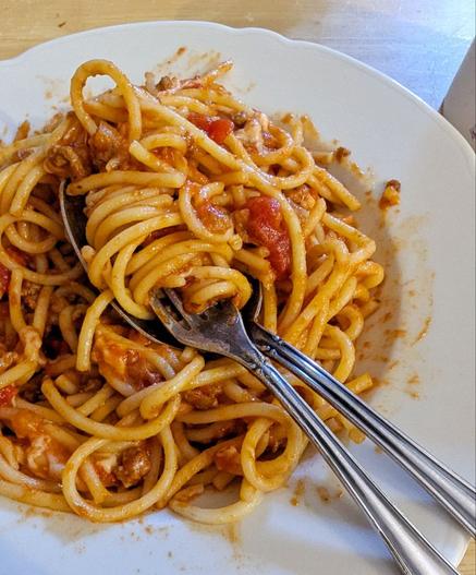 Weißer, tiefer Porzellanteller gefülllt mit Spaghetti in roter Bolognesesauce. Jemand hat schon angefangen zu essen, es sind Saucenspritzer auf dem Teller zu sehen. Gabel und Löffel liegen in der Mitte in einem Nudelwirrwarr.