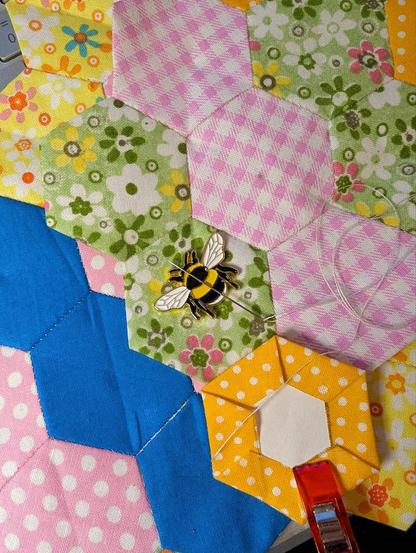 Ein halber Schmetterling aus bunt zusammengesetzten Hexagonformen aus Stoff. Eine Nadel liegt auf einer magnetischen Biene aus Metall (Needle Minder). Darunter liegt ein 2. Schmetterling aus anderen bunten Stoff-Hexies, der schon fertiggestellt ist.