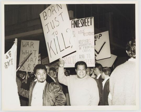 The black-and-white photograph captures a group of individuals participating in what appears to be an anti-government protest. Several people are holding up homemade signs with messages such as "Born just to kill? Finesheet," indicating critical or satirical commentary on the Vietnam War and related societal issues like economic reform, welfare rights, education funding during financial crises, and prison industrial complex expansion.

The expressions of those in the photo range from serious contemplation to smiling engagement. The individuals are dressed casually with jackets, sweaters, and casual shirts indicative of a grassroots or spontaneous gathering rather than an organized event by a specific political party or group. Some faces are partially obscured due to angle and perspective within the crowd's depth.

The setting seems to be indoors under artificial lighting as evidenced by shadows cast on the subjects' bodies and the surrounding architecture not clearly visible, suggesting this might have been taken in a warehouse or large industrial space repurposed for protest activities at that time. The overall composition of the image conveys a sense of activism and collective political expression during a period marked by significant social upheaval related to war involvement and domestic policy challenges.

This photograph is associated with Anti-Government Protest, which could be linked to specific historical period [...]