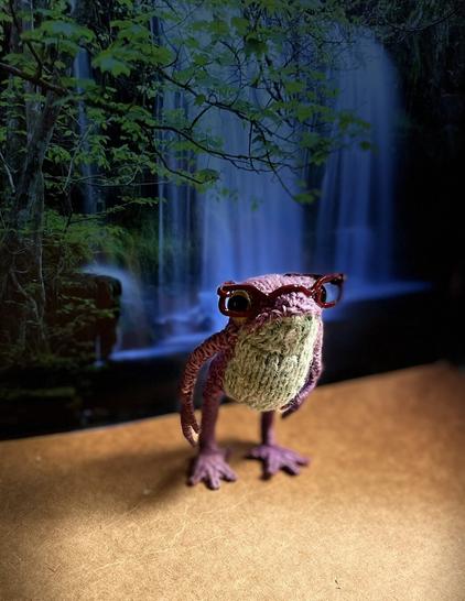 A heliotrope knitted frog with a light turquoise belly stands on a brown surface in front of a waterfall. He’s wearing shiny, dark red glasses, and there’s a spotlight on him.