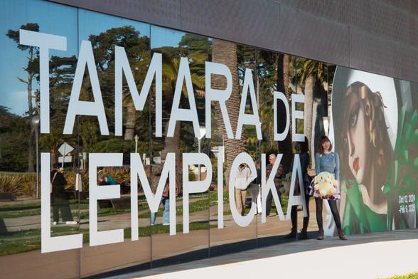 Photo of Lety and the Creature Mastodon stuffed toy standing in front of the Tamara de Lempicka exhibition's window display at de Young Museum. From top to bottom, Lety is seen wearing a tight gray sweater with slits at the shoulder, a floral pink thigh-high skirt, black leggings, and brown boots. She's standing with her back to the dark, ten-foot-tall glass walls of the museum, between “TAMARA DE LEMPICKA” in giant signage and a bust cropping of Lempicka's “Young Woman in Green” with “Oct 12, 2024 - Feb 9, 2025” printed onto the corner. Lety's holding the Creature in front of her lap as she smiles at the camera. Both she and the Creature have a little “Young Woman in Green” sticker on their chests as proof of entry to the exhibit.