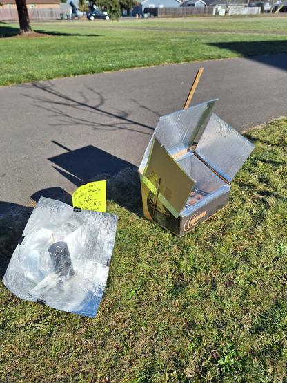 Two different designs for solar cookers, set up in sunny outdoor environment. one is the Copenhagen Cooker panel design, the other is a slant front box oven with 3 reflecting panels. Sign between them reads "these aren't trash, they're SOLAR COOKERS"