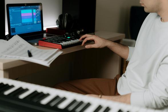 A person in a light-colored long-sleeve shirt and brown pants sits at a wooden desk, working on a music production setup. Their right hand is on a small MIDI keyboard and drum pad controller with red and black accents. A laptop screen displays a digital audio workstation (DAW) with multiple audio tracks. A pair of headphones rests on the desk. In the foreground, a full-sized keyboard piano is partially visible. Scattered sheet music with a pen lies on the desk, indicating active composition or arrangement work.