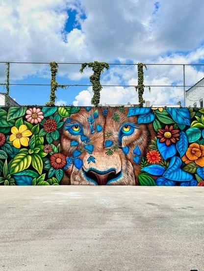Streetartwall. A beautiful mural of a lion's head peeking out from colorful flowers was painted on a long wall in a Brooklyn school. The photo shows just a section of a long wall in the schoolyard, with different colored flowers, leaves and vines, in the middle of which a friendly-looking lion's head appears. Simply wonderful colors and great painting.
