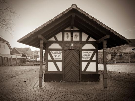 Das Backhaus in Münster im Landkreis Gießen. Die Aufnahme in Sepia-Tönung verleiht dem Bild eine nostalgische und zeitlose Atmosphäre. Im Hintergrund sind ländliche Häuser und Zäune zu sehen, die die dörfliche Umgebung unterstreichen. Das Foto vermittelt eine ruhige, geschichtsträchtige Stimmung.
